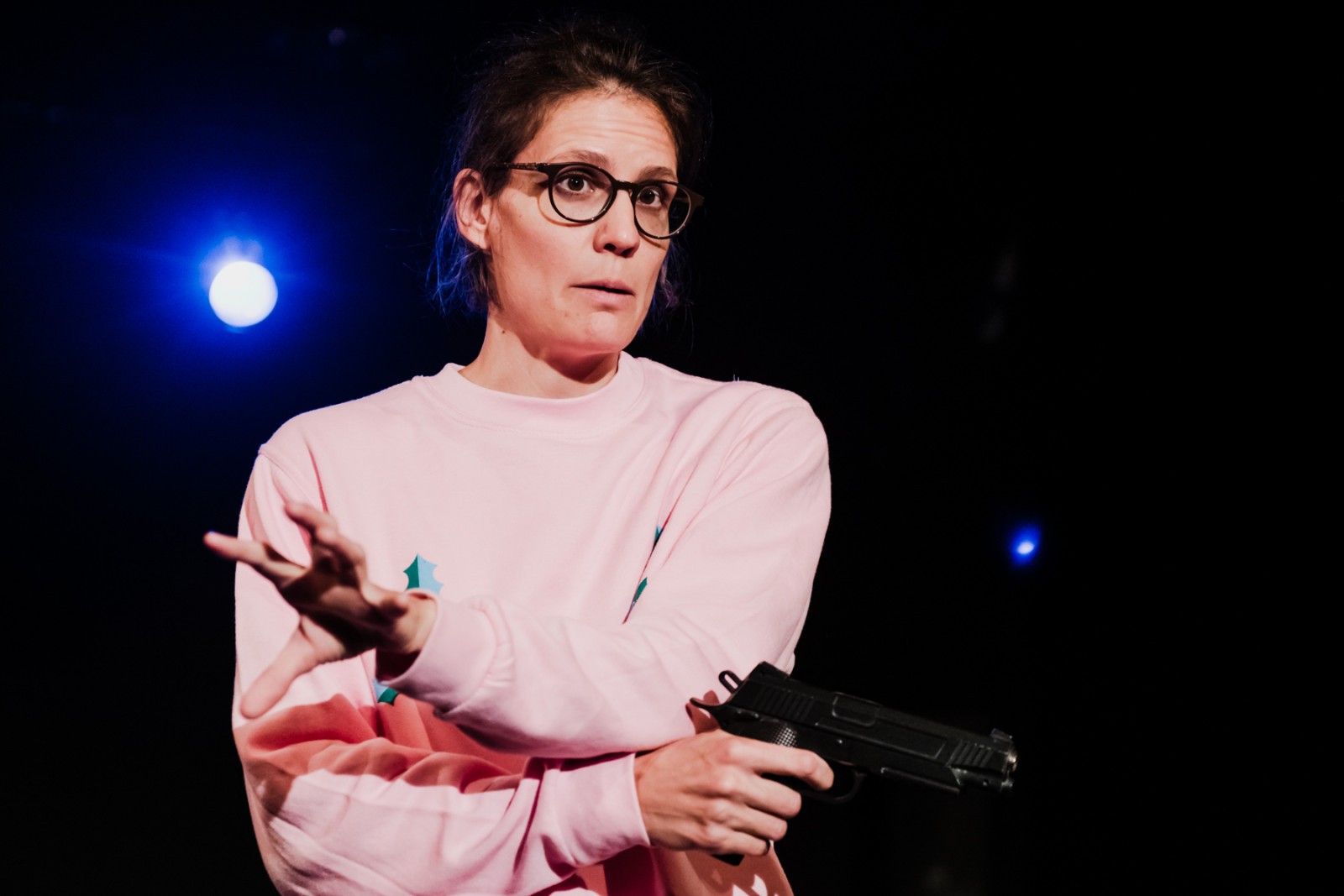 Eine Frau mit braunen Haaren und einer Brille schaut erwartungsvoll ins Publikum. Sie hält in einer Hand eine Schusswaffe mit der anderen Hand gestikuliert sie.