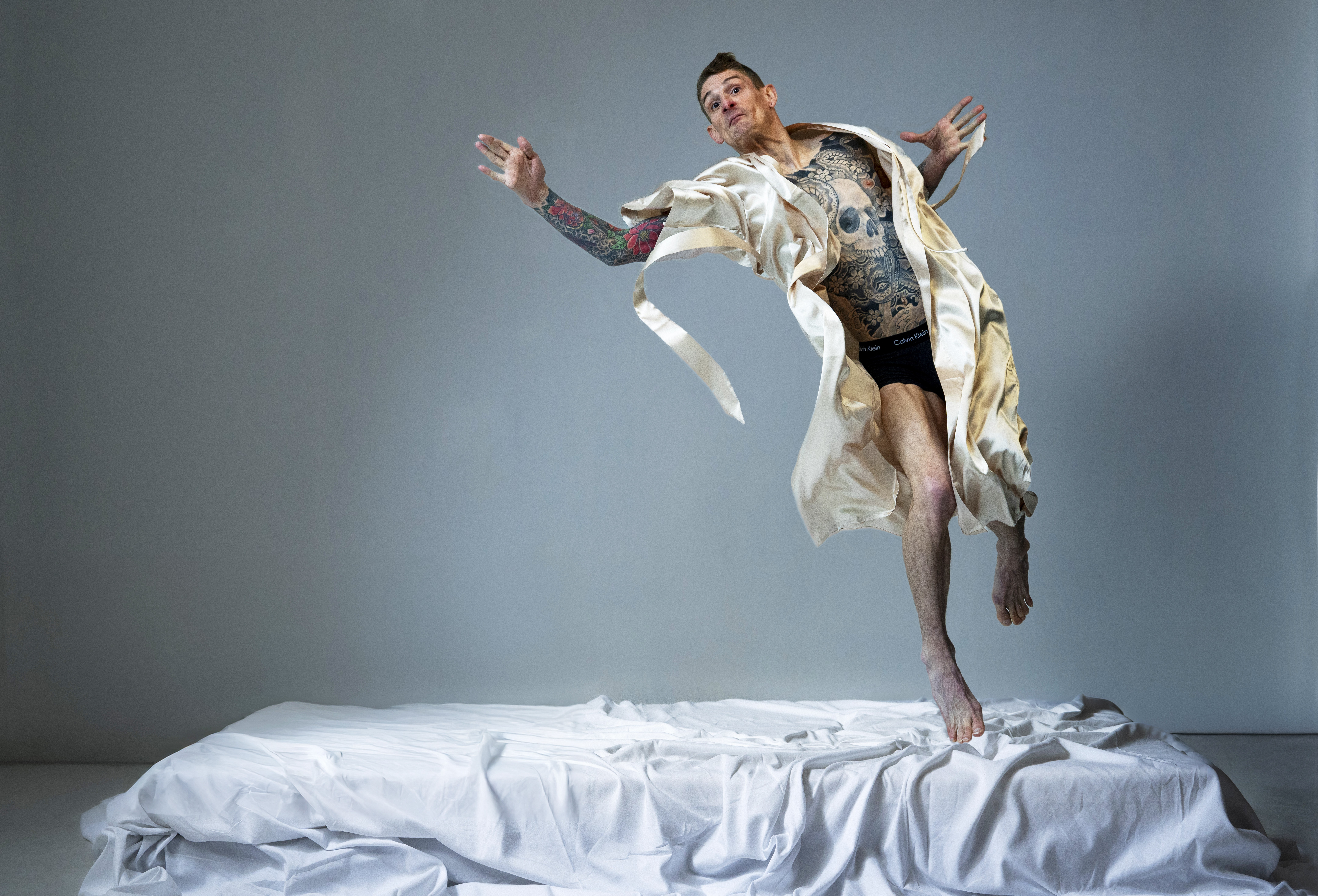 Ein Performer springt in die Luft und schaut dabei in die Kamera. Er trägt eine Unterhose und einen goldenen Bademantel. Sein Körper ist tätowiert.