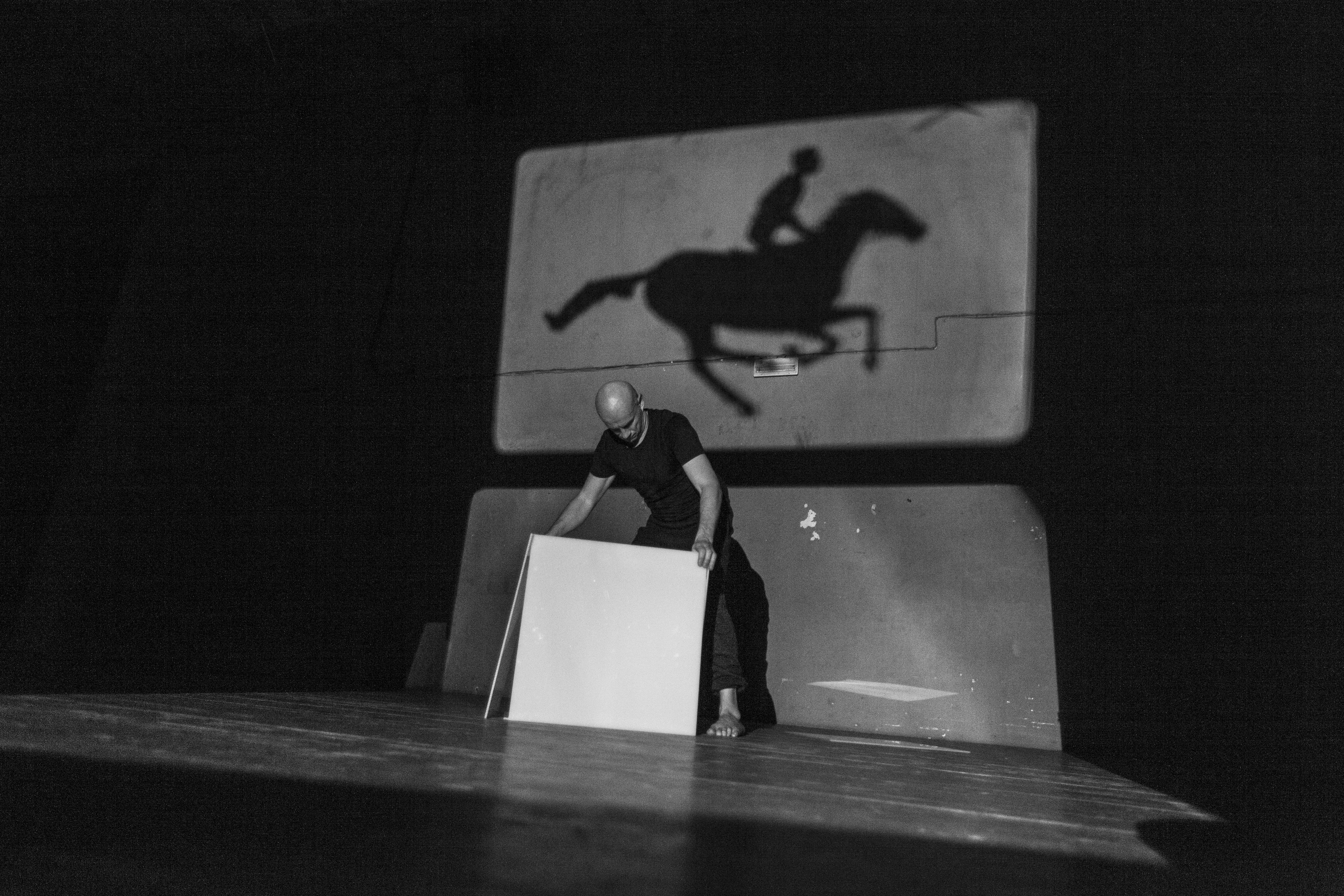 Schwarzweiss Bild mit der Schattenfigur eines galopierenden Pferdes mit Reiter*in im Hintergrund. Im Vordergrund der Künstler während seiner Performance.