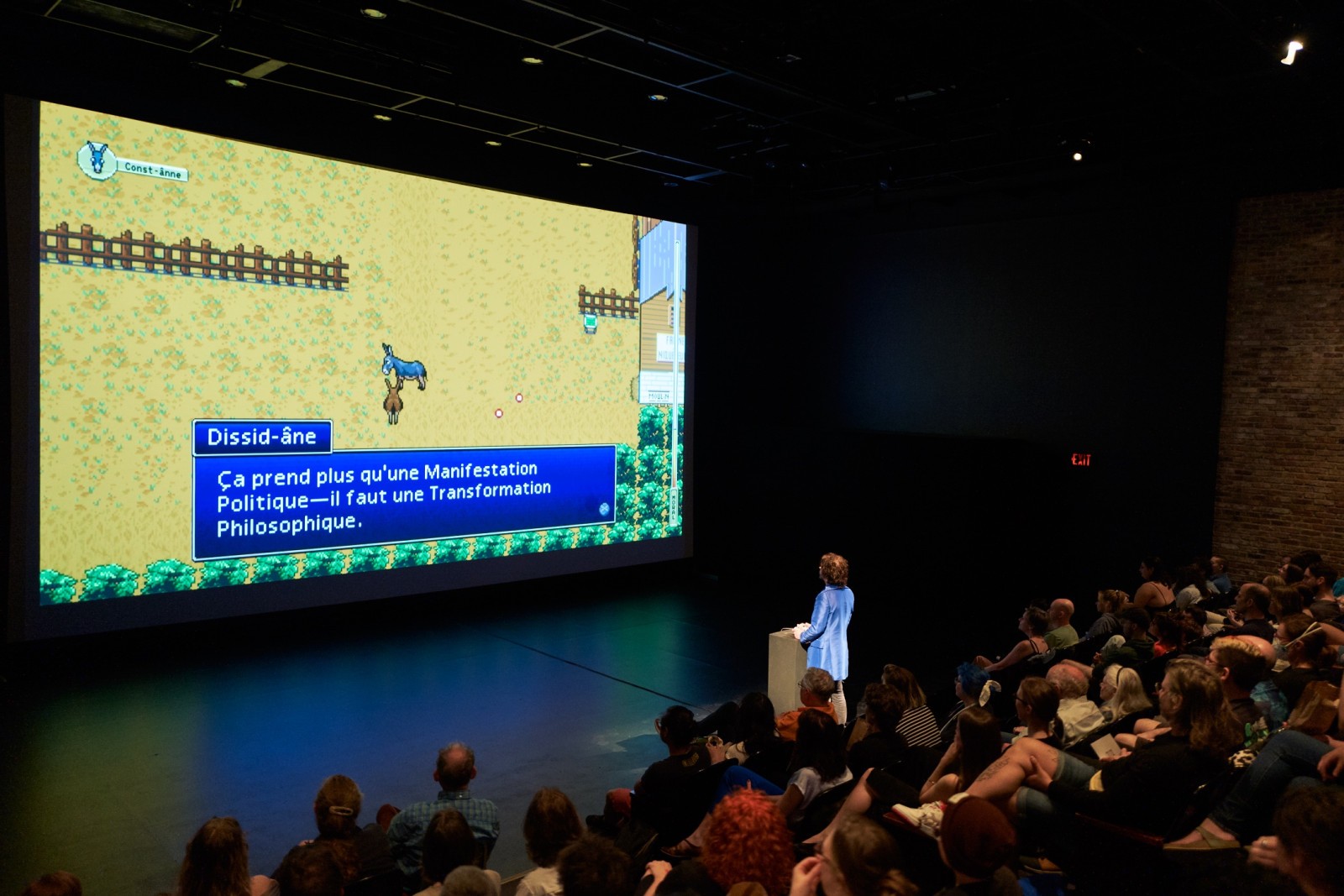 Auf der rechten Seite des Bildes sieht man das Publikum sitzen, auf der linken Seite ist eine grosse Leinwand auf der ein Videospiel mit zwei Esel projiziert ist. Eine Person steht vor dem Publikum und schaut Richtung Leinwand.