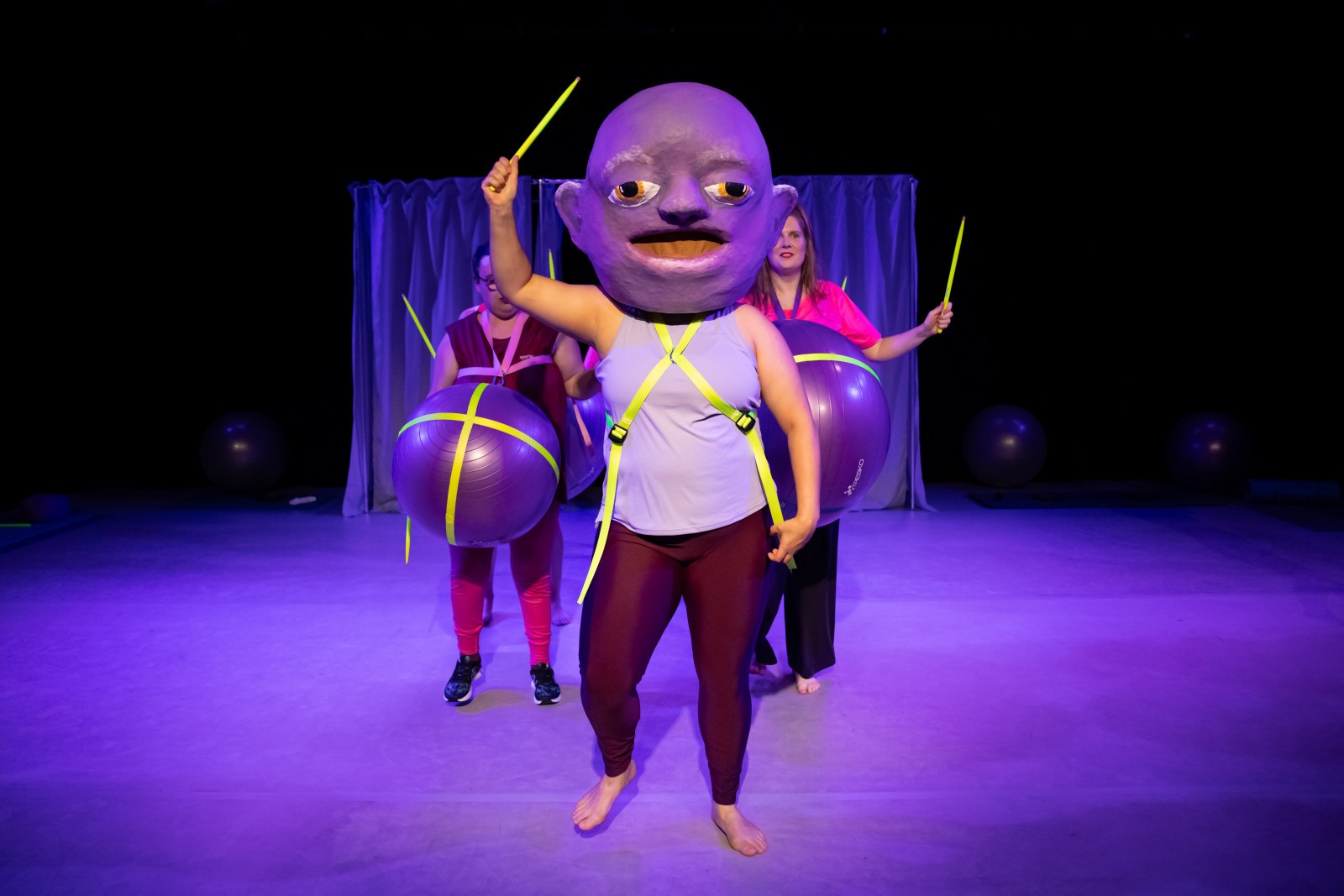 Drei Menschen stehen auf der Bühne. Eine Person trägt einen übergrossen Kopf als Maske, die anderen Beiden tragen violette Gymnastikbälle vor ihren Bäuchen.