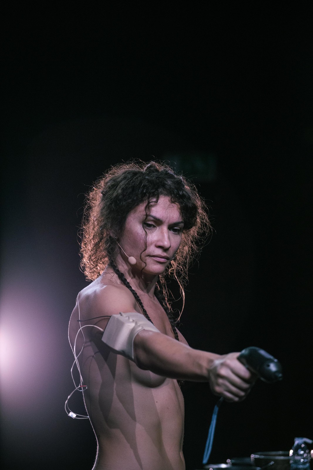 Eine Frau steht seitlich und hält einen schwarzen Gegenstand wie eine Schusswaffe in der Hand. Sie trägt keine Kleidung und schaut ernst zu Boden.