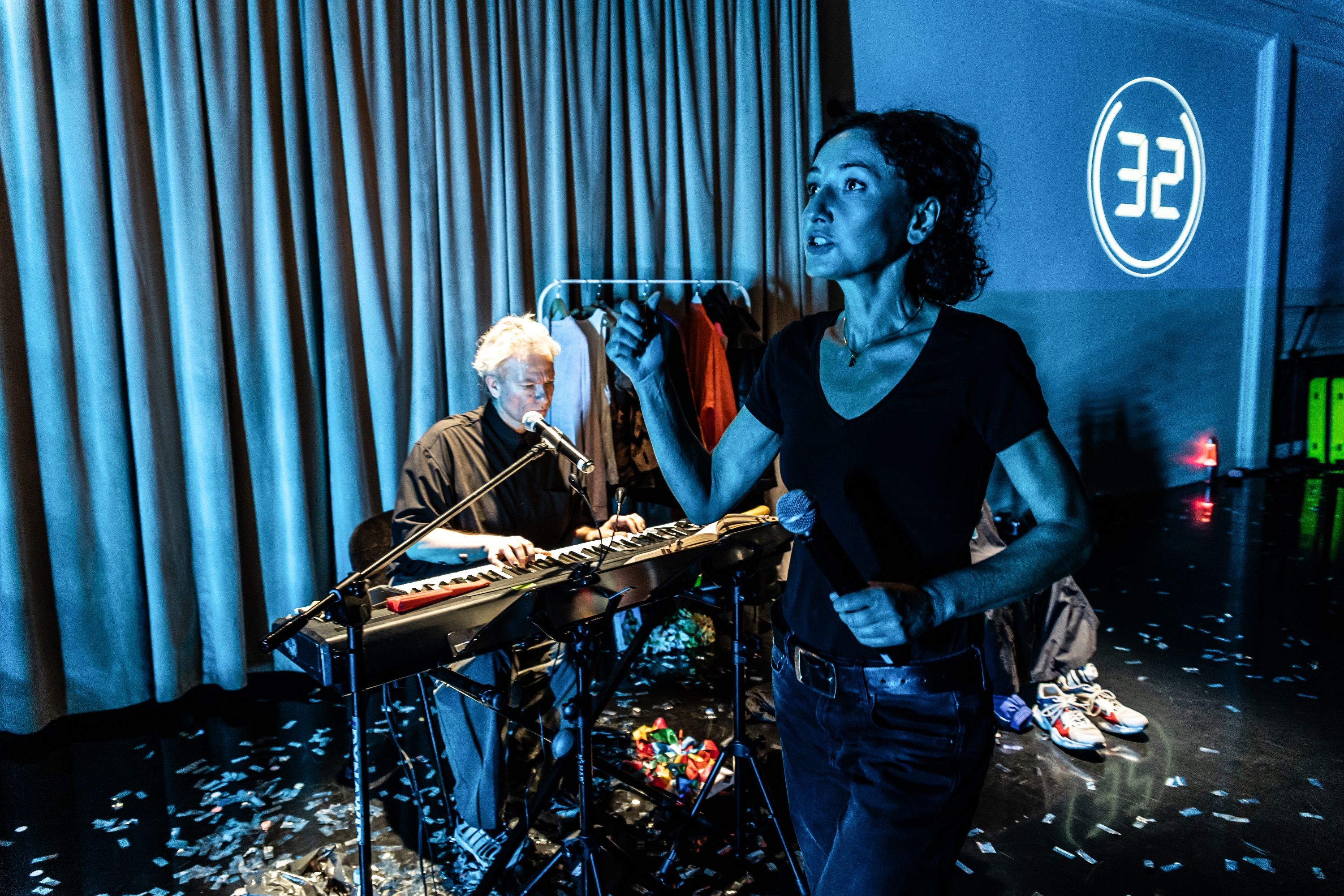 Ein Performer sitzt an einem Key Board. Eine Performerin hält ein Mikrofon in der Hand und ist in Bewegung. Auf dem Boden liegen Konfetti.