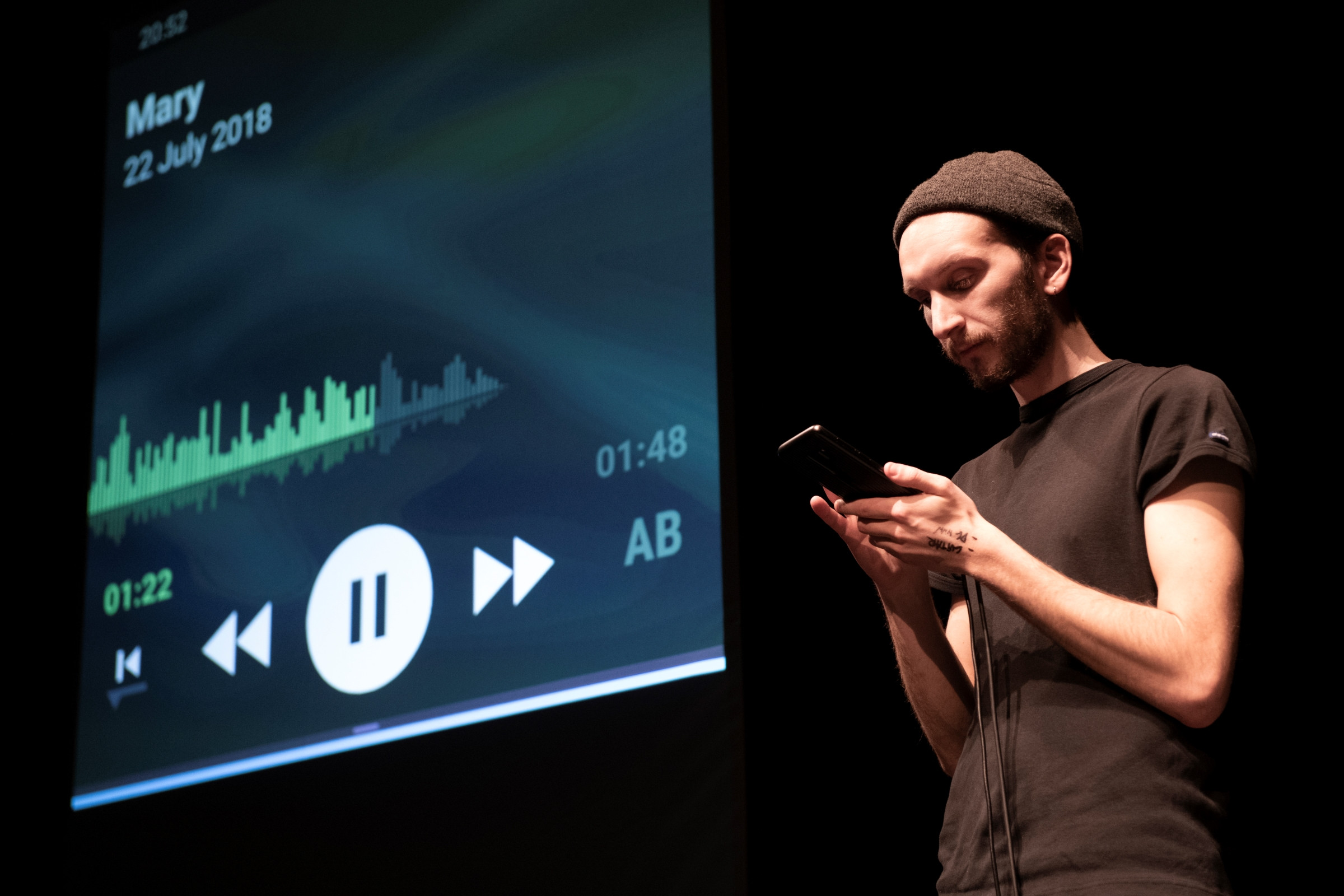 Hannes Dereere mit Blick auf das Smartphone in seiner Handy. Der Screen neben ihm zeigt das Abspielen einer Tonaufnahme.