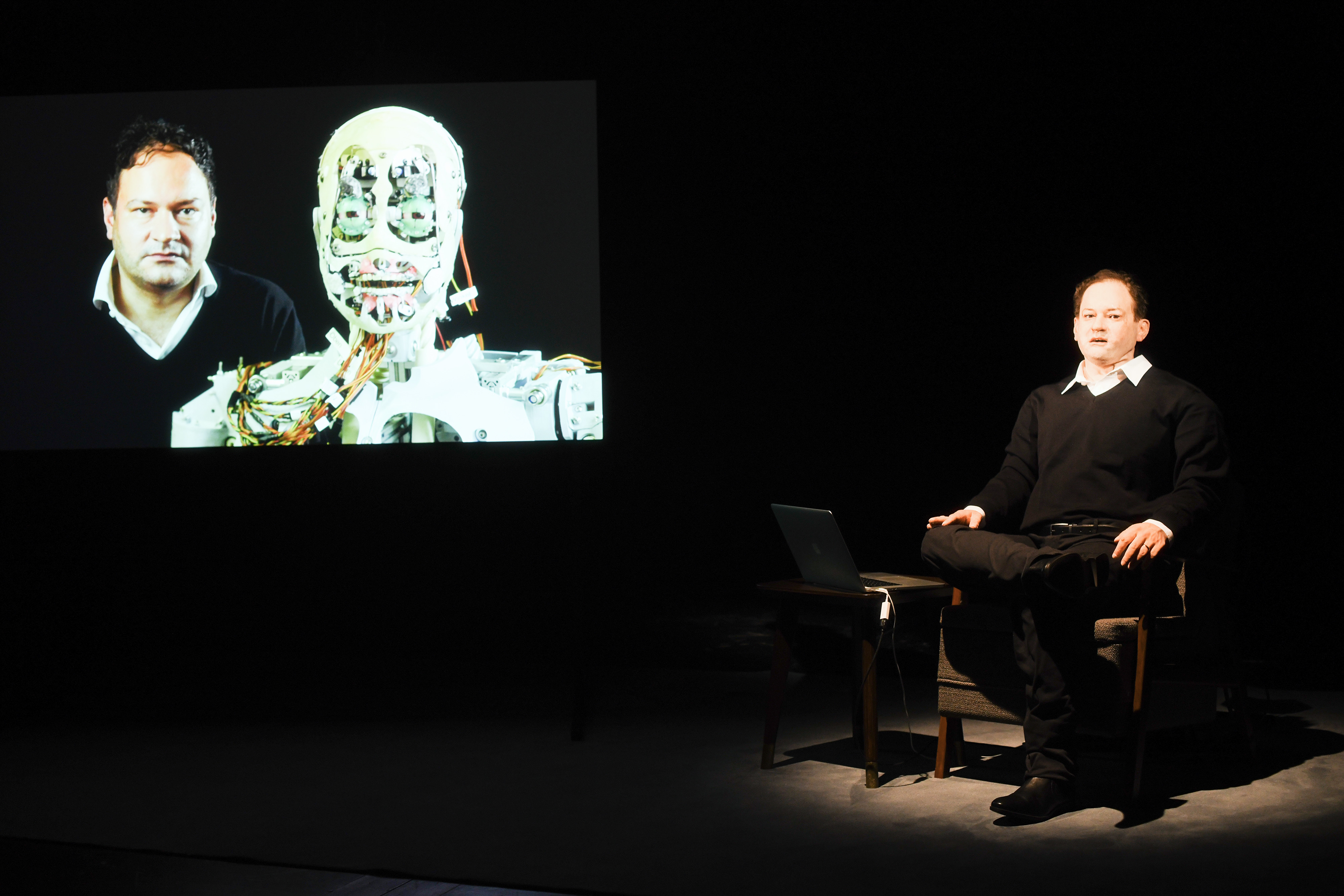Links im Bild ist ein Bildschirm, auf welchem ein Portrait eines Mannes und der Technik des Roboterkopfes zu sehen ist. Links sitzt der Roboter mit dem Gesicht dieses Mannes.
