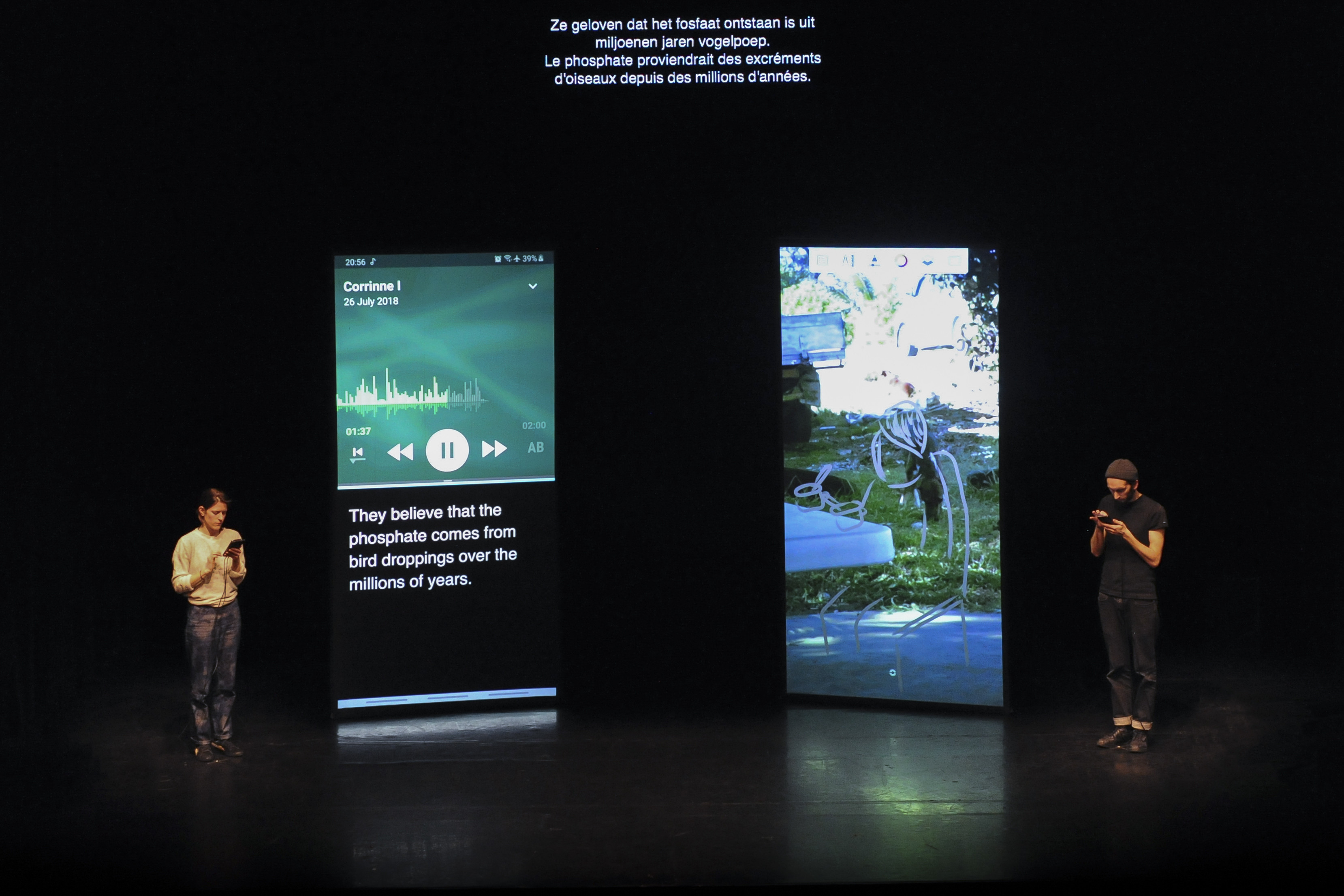Die beiden Künstler*innen stehen neben grossen Screens, die die Smartphones in den Händen der beiden zeigen.