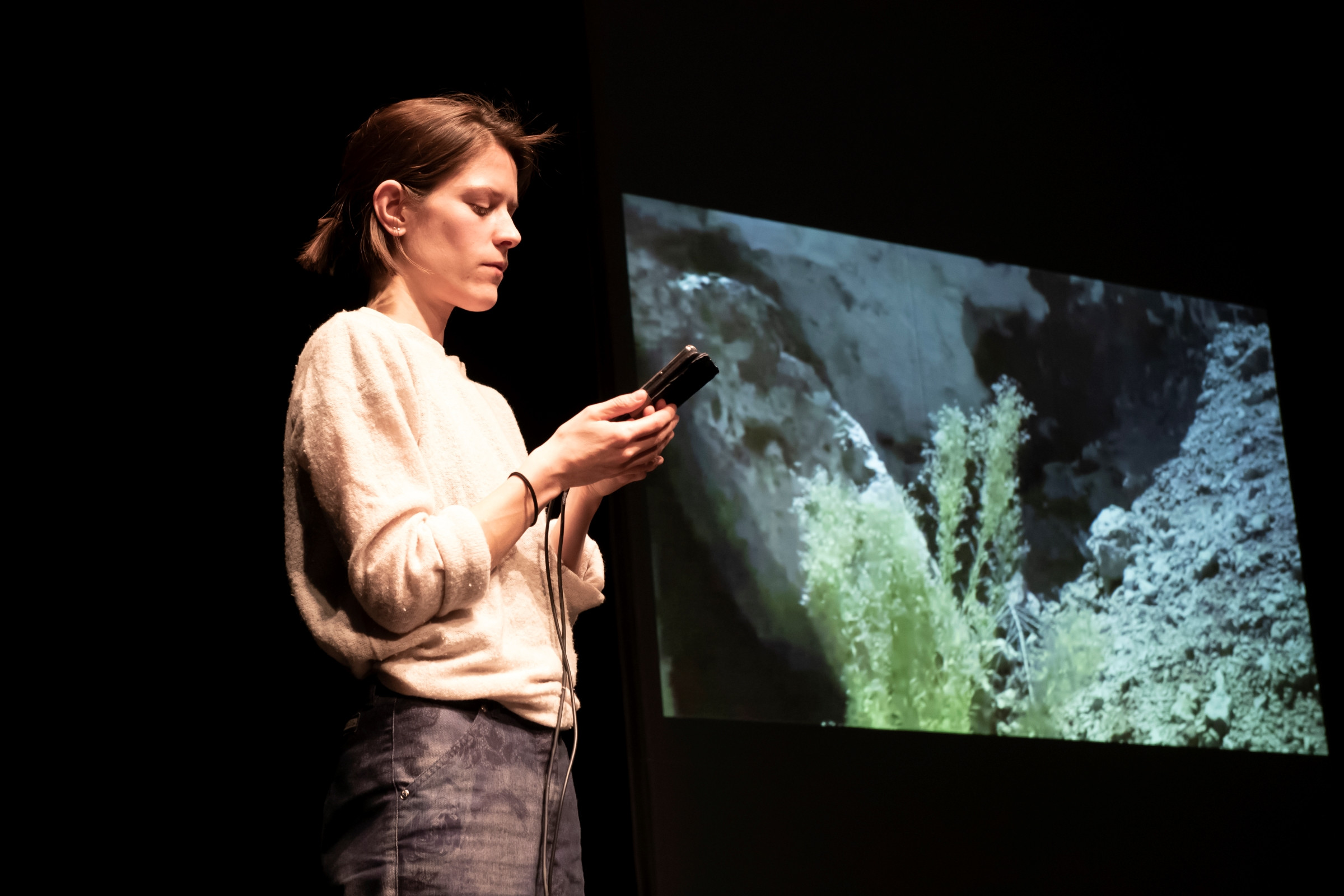 Silke Huysmans mit Blick auf das Smartphone in ihrer Hand. Im Hintergrund ein Foto auf einem Screen.